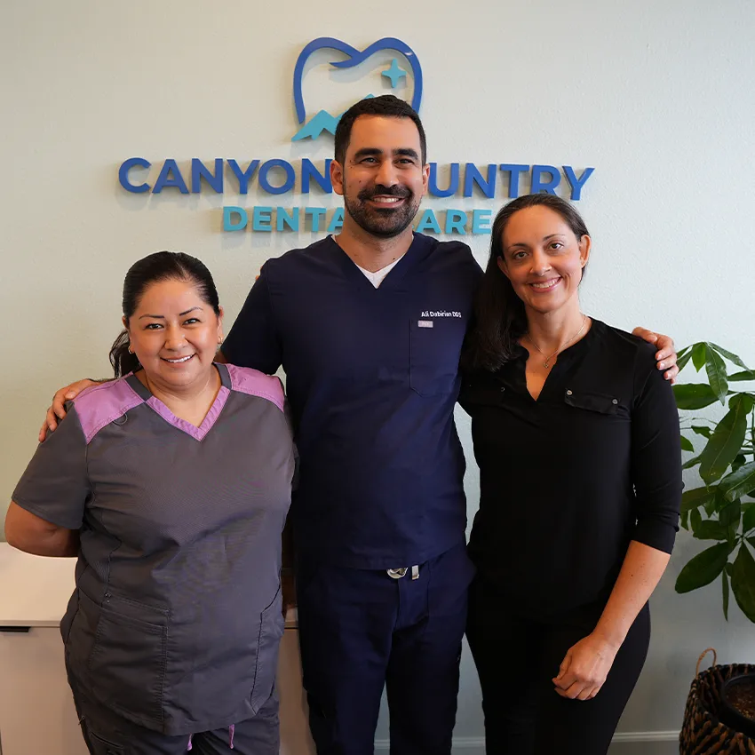 Dr. Dabirian and two members of the Canyon Country Dental Care team standing in front of the office logo wall