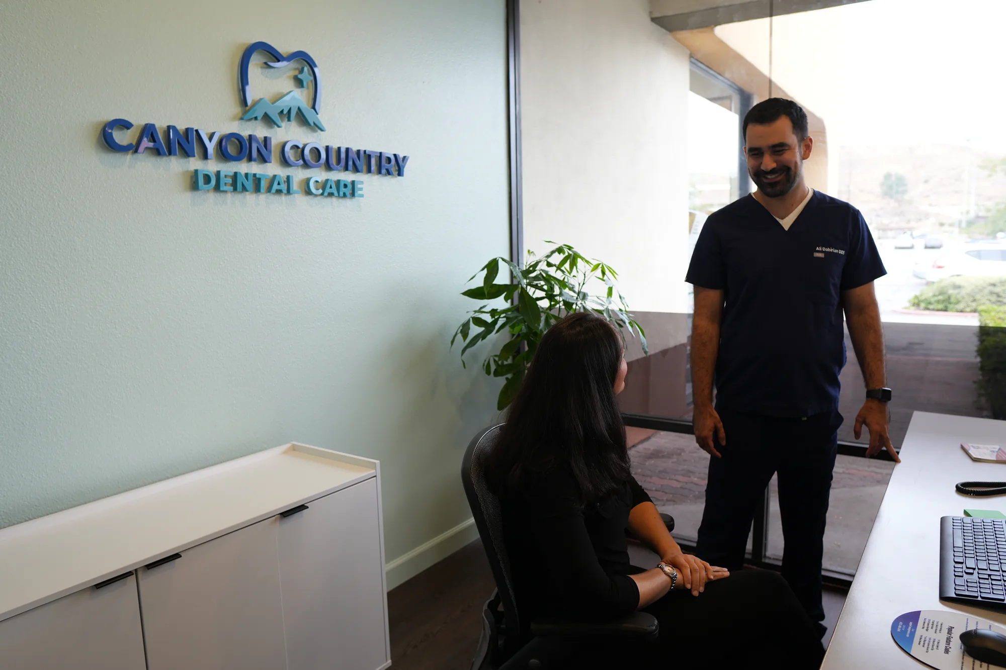 Dr. Dabirian standing beside a woman seated at the front desk computer, in front of the logo wall