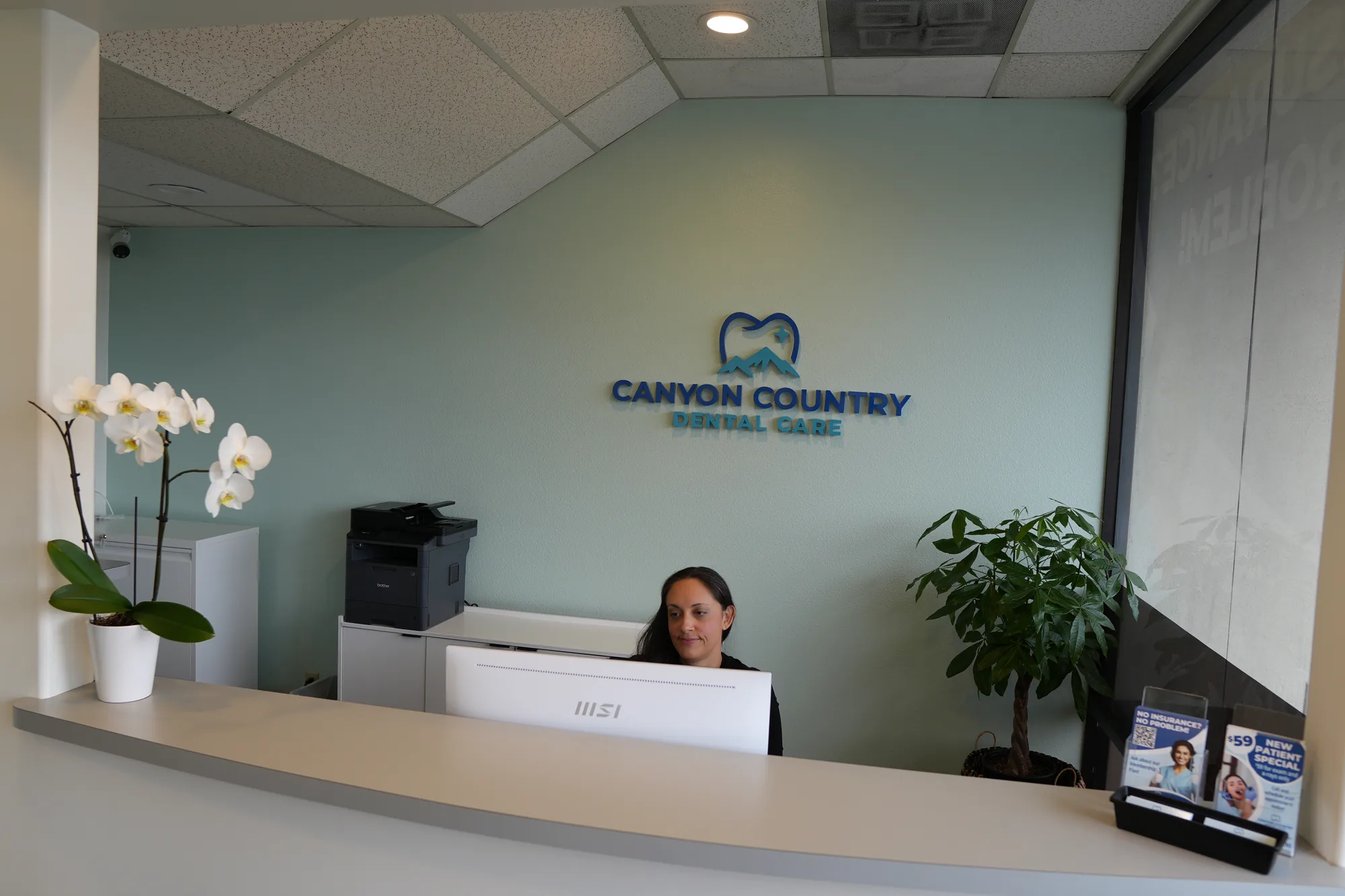 Reception desk with a person seated behind it, orchids on the counter and the Canyon Country Dental Care logo on the wall