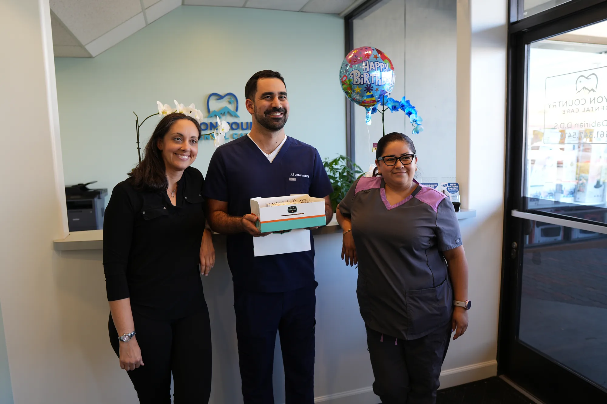 Dr. Dabirian and two team members at the reception counter, holding a box, with a balloon behind them