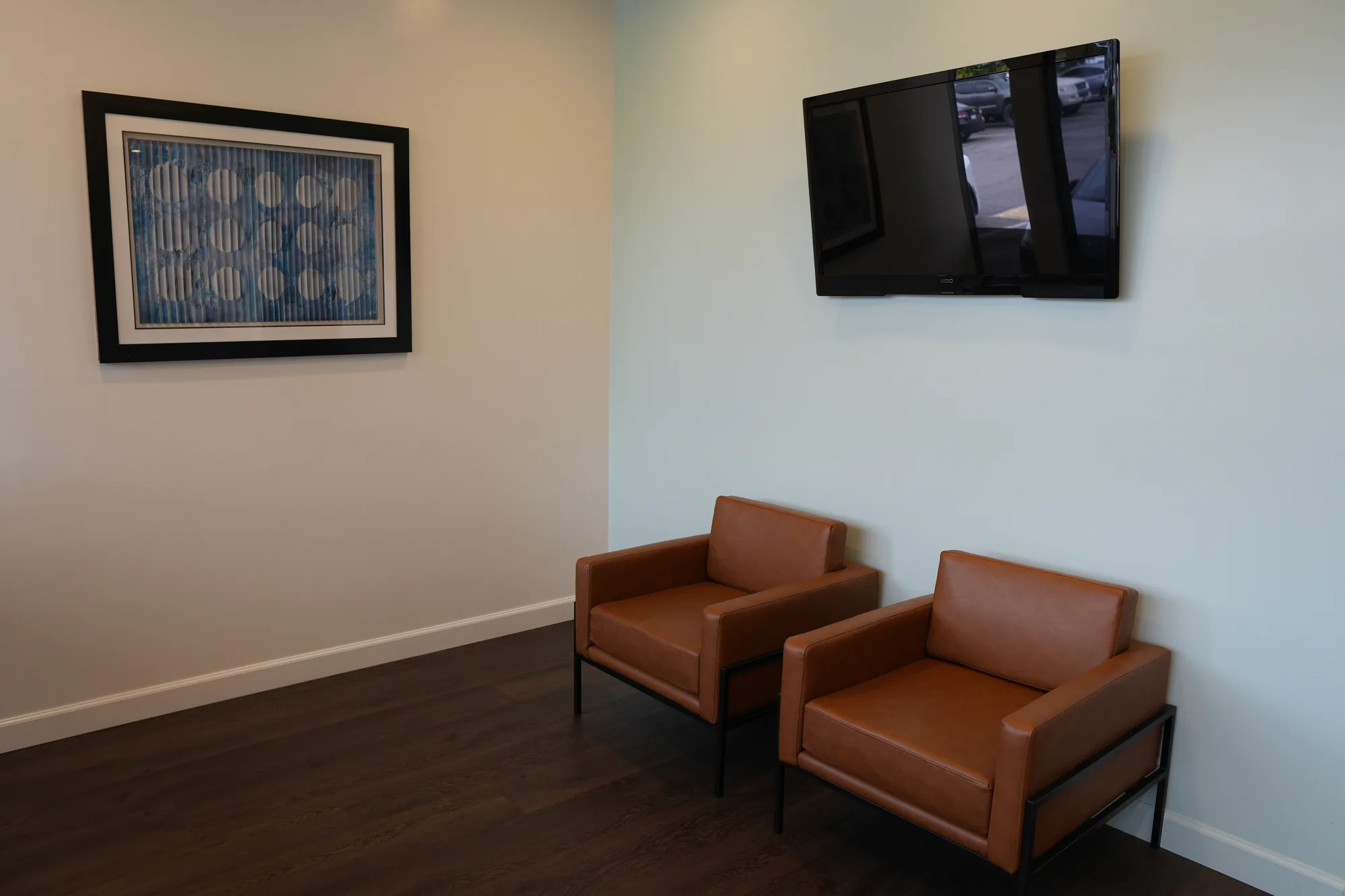 Two brown leather armchairs in the waiting room, with a framed picture and a wall-mounted TV